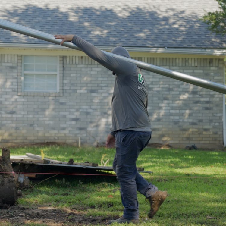 Worker carrying long steel pipe across yard by brick house