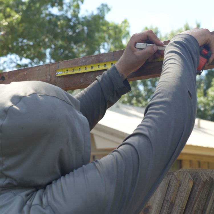 Screenshot 2025-10-07 at 4.12.56 PM (Custom) Worker in hood measuring top rail of wood fence with tape